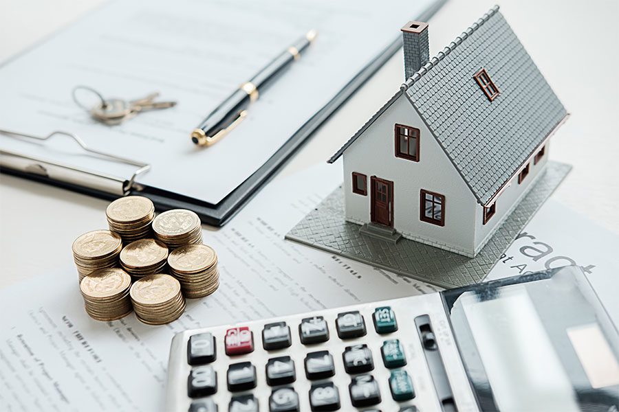 Home Warranty - View of a Model House Sitting on a Table on Top of a Mortgage Application Next to a Stack of Coins a Calculator and a Notepad with Keys and a Contract