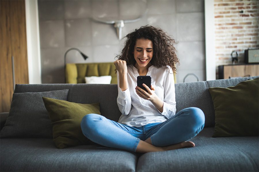 Resources - Closeup Portrait of a Cheerful Young Woman Sitting on the Sofa at Home Celebrating Success While Using Her Phone