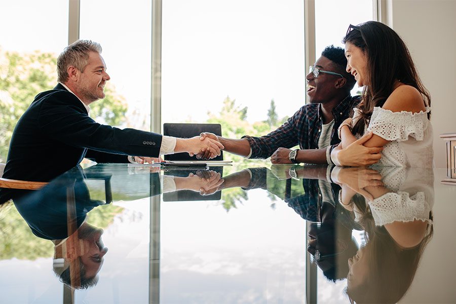 About Our Agency - View of a Realtor Sitting in a Modern Office with a Young Couple Shaking Their Hands to Celebrate Success in Finding a New Home