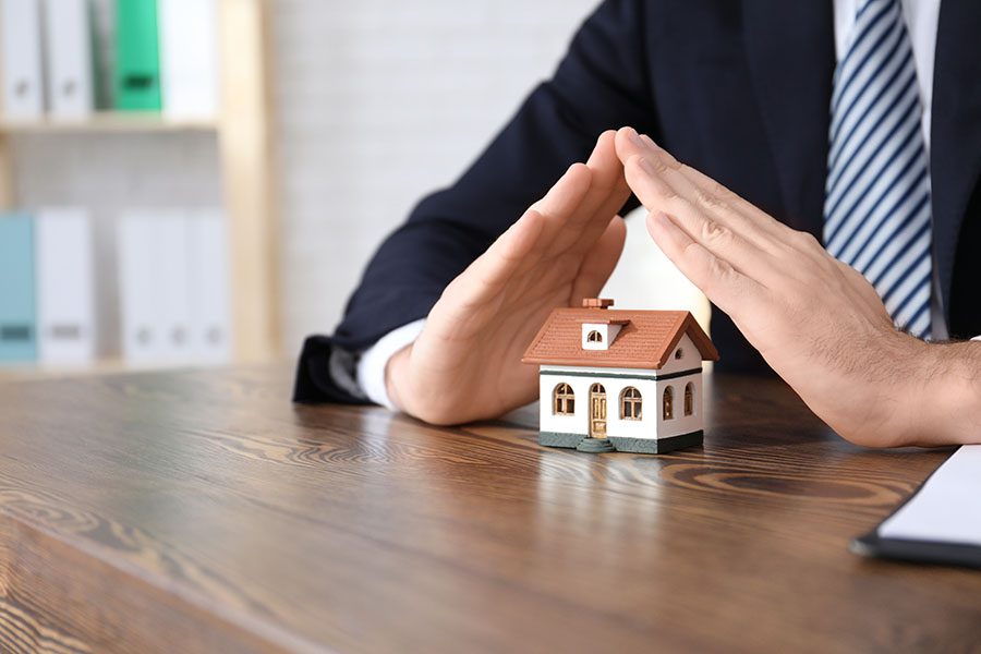 Direct to Consumer Home Warranty - Closeup Portrait of an Insurance Agent Sitting in an Office Holding His Hands Over a Small Toy Model House